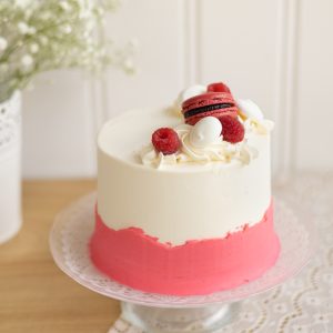 image du gâteau d'anniversaire vanille-framboise : Gâteau à la vanille garni de chantilly à la framboise, masqué de crème au beurre à la vanille avec une bordure rose. Le décor est composé de macaron à la framboise, de meringues, de framboise fraîche et de perle nonpareils blanches.