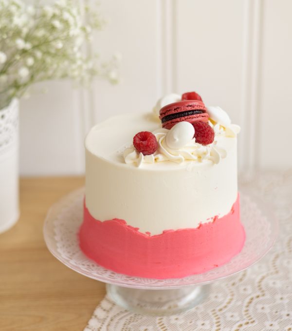 image du gâteau d'anniversaire vanille-framboise : Gâteau à la vanille garni de chantilly à la framboise, masqué de crème au beurre à la vanille avec une bordure rose. Le décor est composé de macaron à la framboise, de meringues, de framboise fraîche et de perle nonpareils blanches.