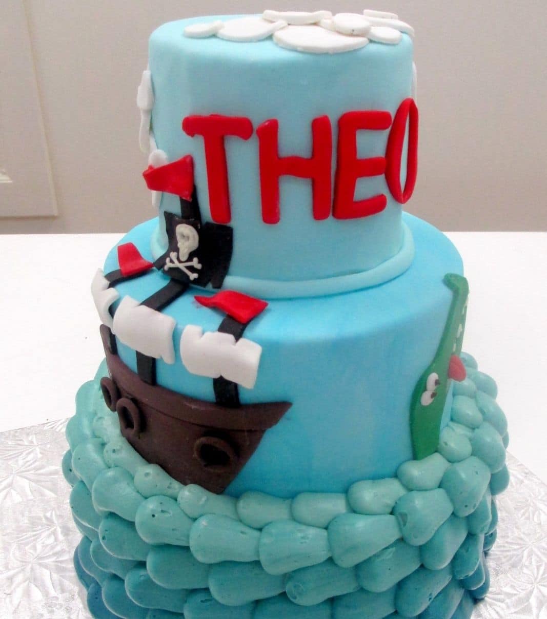 Gâteau d'anniversaire sur mesure de Théo: gâteau de 3 étages à thématique Peter Pan avec la base en couverture à la crème au beurre dégradée de bleu à turquoise, le 2e et 3e étage avec une couverture en fondant et décoré d'un bateau de pirate et d'un crocodile 2D en fondant.