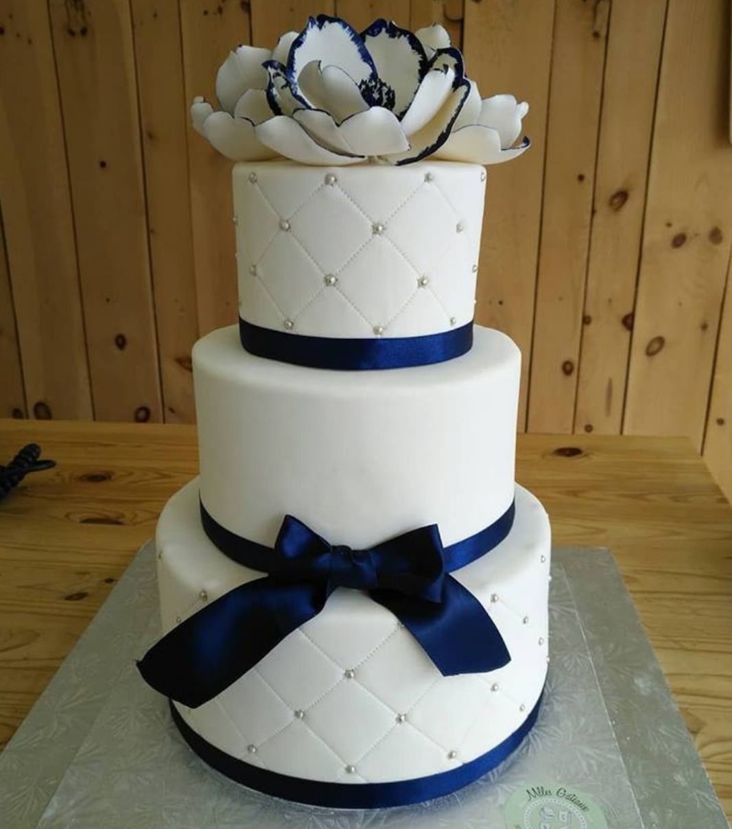Gâteau de mariage: gâteau de 3 étages à thème blanc et marine, avec une couverture fondante à effet matelassé (quilting) décoré de perles argentées, de fleurs en sucre et de rubans de tissu marine.