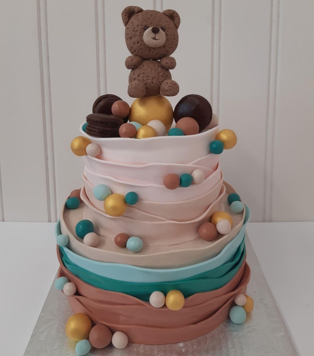 Gâteau shower de bébé: gâteau de 2 étages avec une couverture au fondant texturée en bandelettes dégradées de couleurs et décoré d'une figurine d'ourson, de macarons et de boules au fondant roses, dorées, terracotta et turquoises.