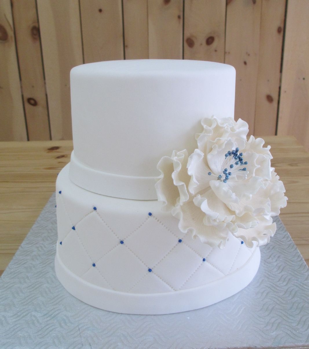 Gâteau d'anniversaire sur mesure: gâteau de 2 étages avec une couverture en fondant à effet matelassé (quilting) avec des points bleus et décoré d'une grosse fleur en sucre agrémentée de détails bleus.