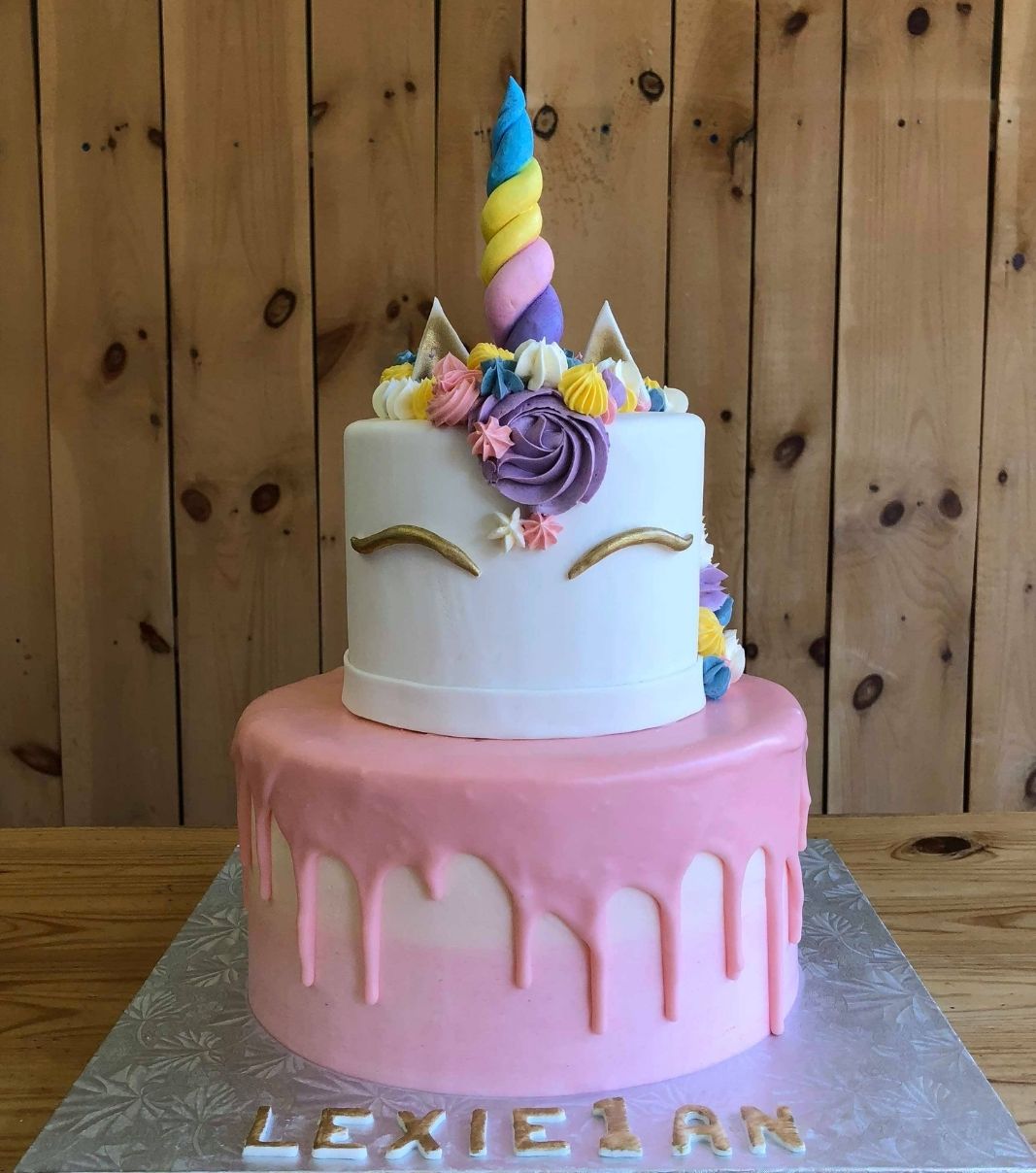 Gâteau d'anniversaire sur mesure du 1 an de Lexie: gâteau de 2 étages à thématique de licorne avec un coulis rose sur une couverture en fondant et décoré d'une corne et d'oreilles en fondant et d'une crinière en crème au beurre multicolore.