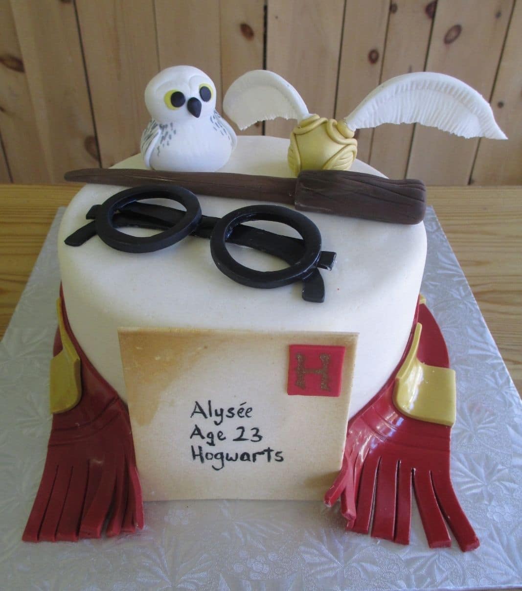 Gâteau d'anniversaire sur mesure des 23 ans d'Alysée: gâteau à thématique Harry Potter avec une couverture en fondant et décoré avec un hibou Hedwige, un vif d'or, un foulard Gryffondor, une baguette magique, une paire de lunettes et une lettre d'invitation à Poudlard en fondant.