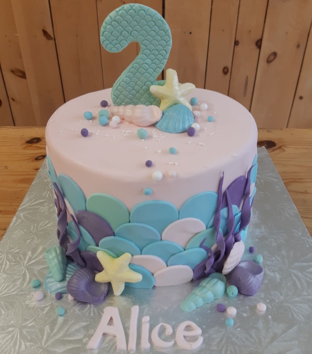 Gâteau d'anniversaire sur mesure des 2 ans d'Alice: gâteau à thématique de sirène avec une couverture en fondant et décoré d'items de la mer, de coquillages et du chiffre 2 couvert d'écailles en fondant.