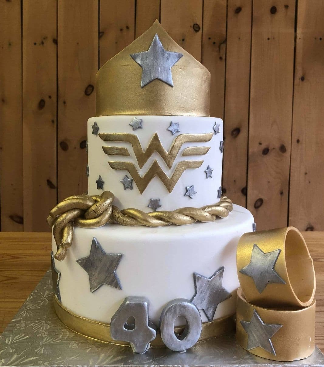 Gâteau d'anniversaire sur mesure de 40 ans: gâteau de 2 étages à thématique Wonder Woman avec une couverture en fondant blanc, doré et argent et décoré d'une couronne, d'étoiles et de bracelets en fondant.