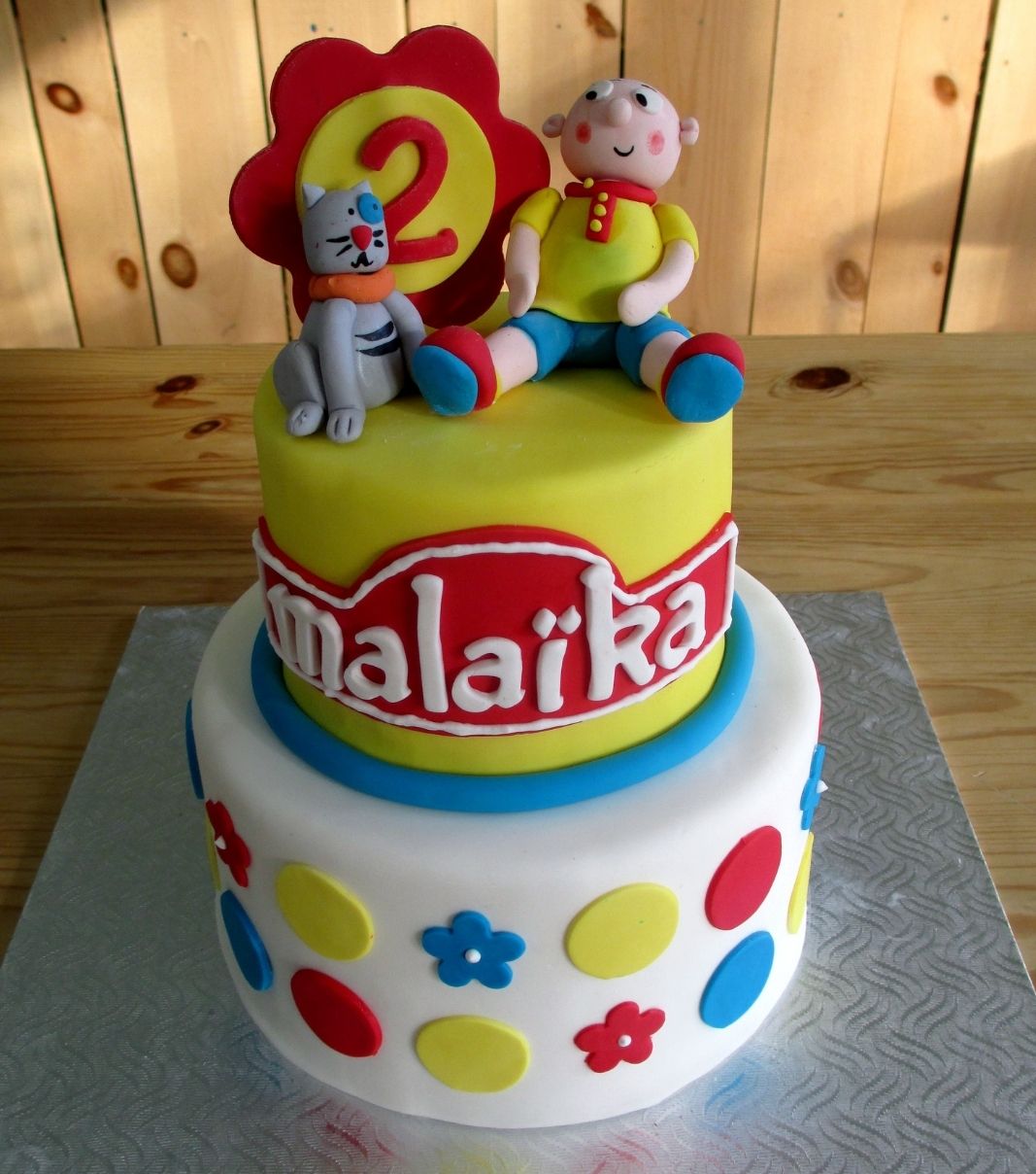Gâteau d'anniversaire sur mesure des 2 ans de Malaïka: gâteau de 2 étages à thématique Caillou et son chat Gilbert avec une couverture en fondant blanc et décoré de cercles de couleur et de fleurs en fondant.