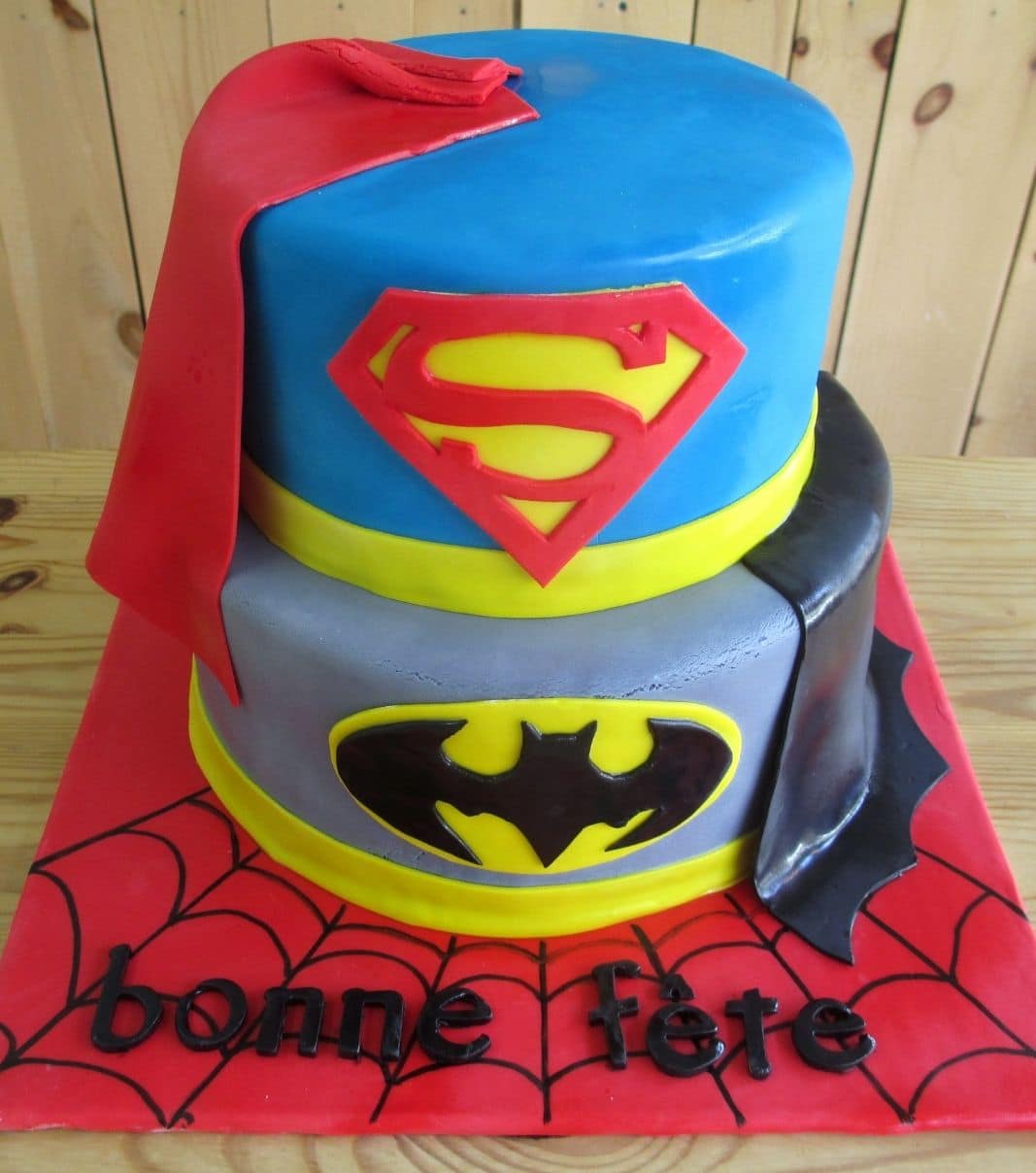 Gâteau d'anniversaire sur mesure: gâteau de 2 étages à thématique Superhéros avec une couverture en fondant, avec la base à thème de Batman, le 2 étage à thème de Superman, avec le carton décoré avec des toiles d'araignée de Spiderman et décoré de capes en fondant.