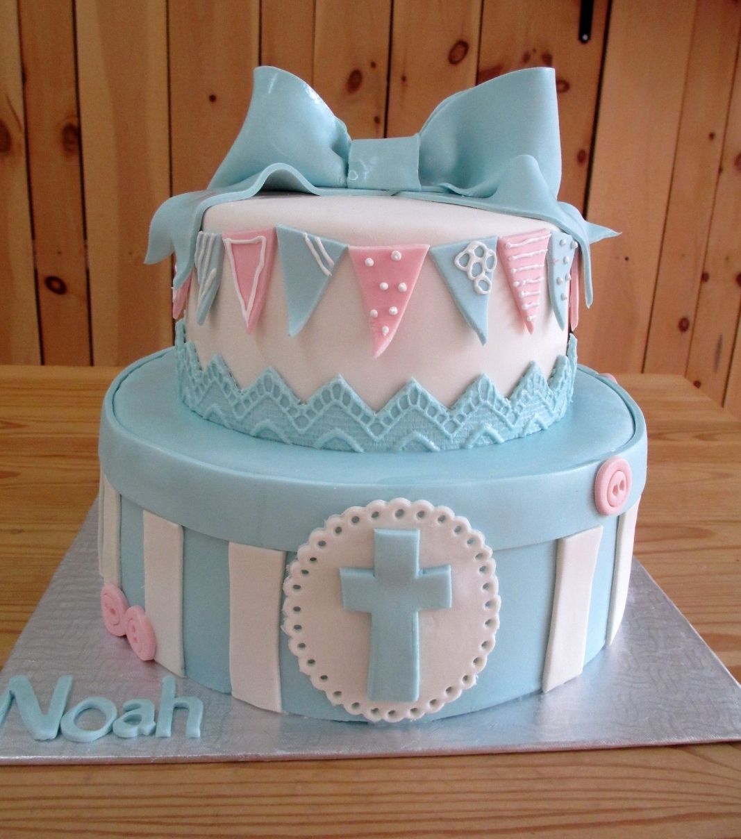 Gâteau de baptême de Noah: gâteau de 2 étages avec une couverture en fondant rose et bleu et décoré d'une boucle et de dentelle bleues, de boutons roses et de fanions.