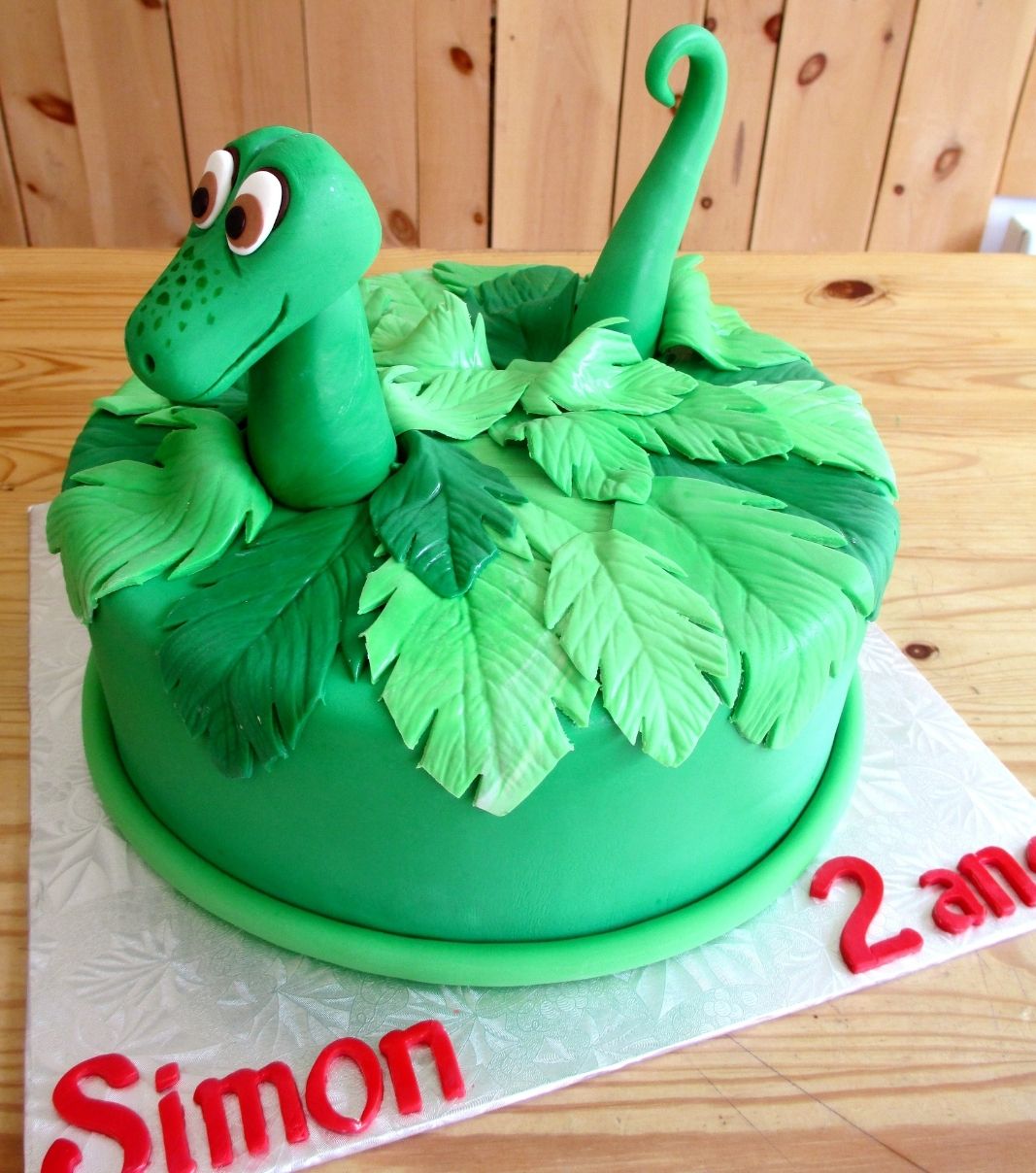 Gâteau d'anniversaire sur mesure des 2 ans de Simon: gâteau à thématique de dinosaure avec un feuillage de fougères, une tête et une queue de dinosaure en fondant.