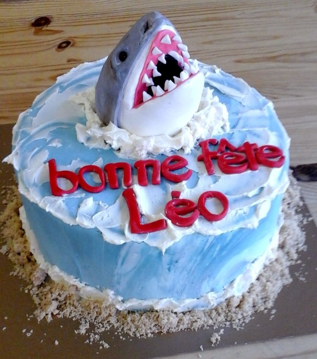 Gâteau d'anniversaire sur mesure des 5 ans de Léo: gâteau à thématique de requin avec une couverture texture eau de la mer et une bordure de sable et décoré avec une figurine de tête de requin en fondant.
