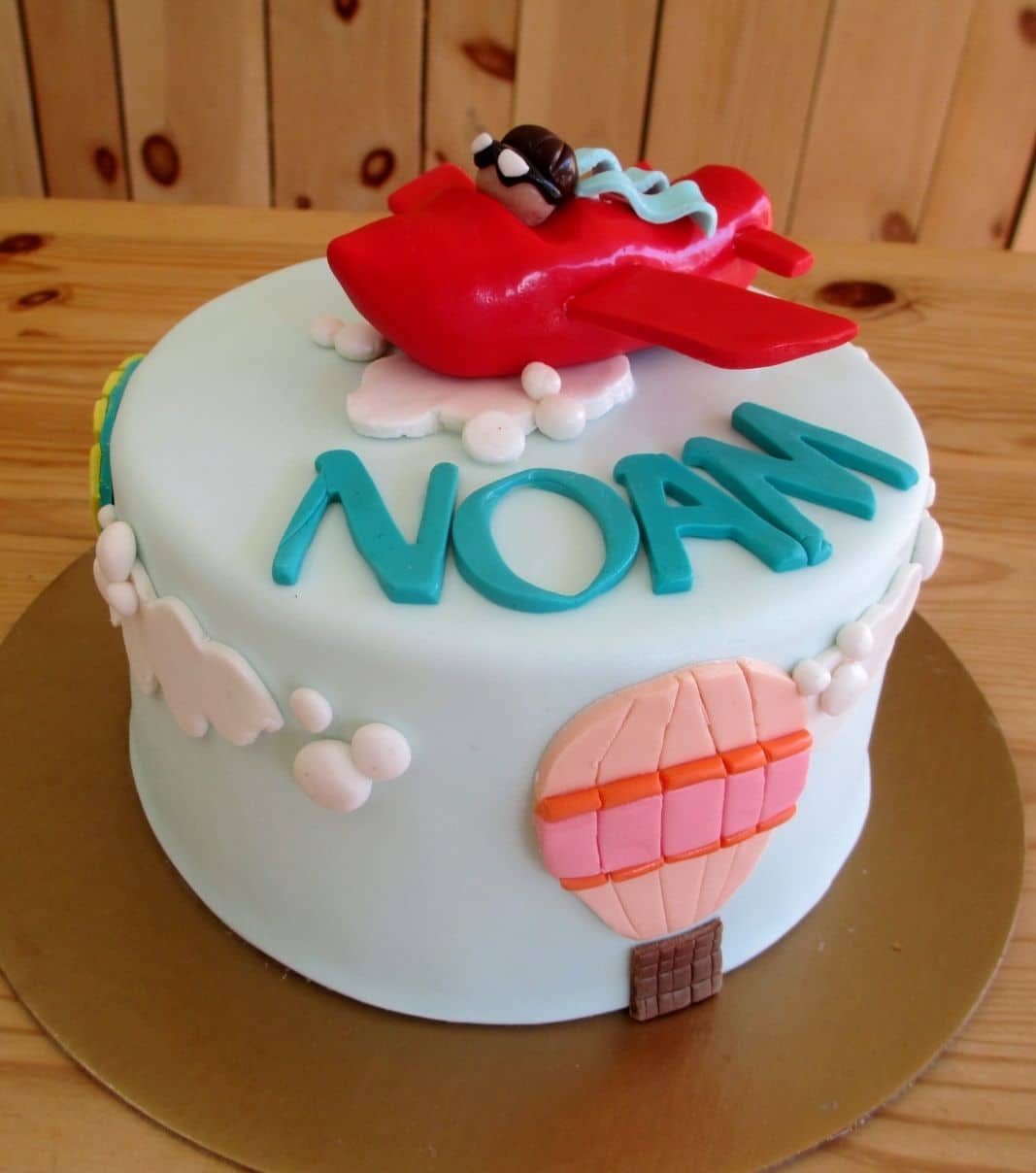 Gâteau d'anniversaire sur mesure de Noam: gâteau à thématique d'avion et montgolfière avec une couverture en fondant en décoré d'un avion avec son pilote, de nuages et de montgolfières 2D en fondant.
