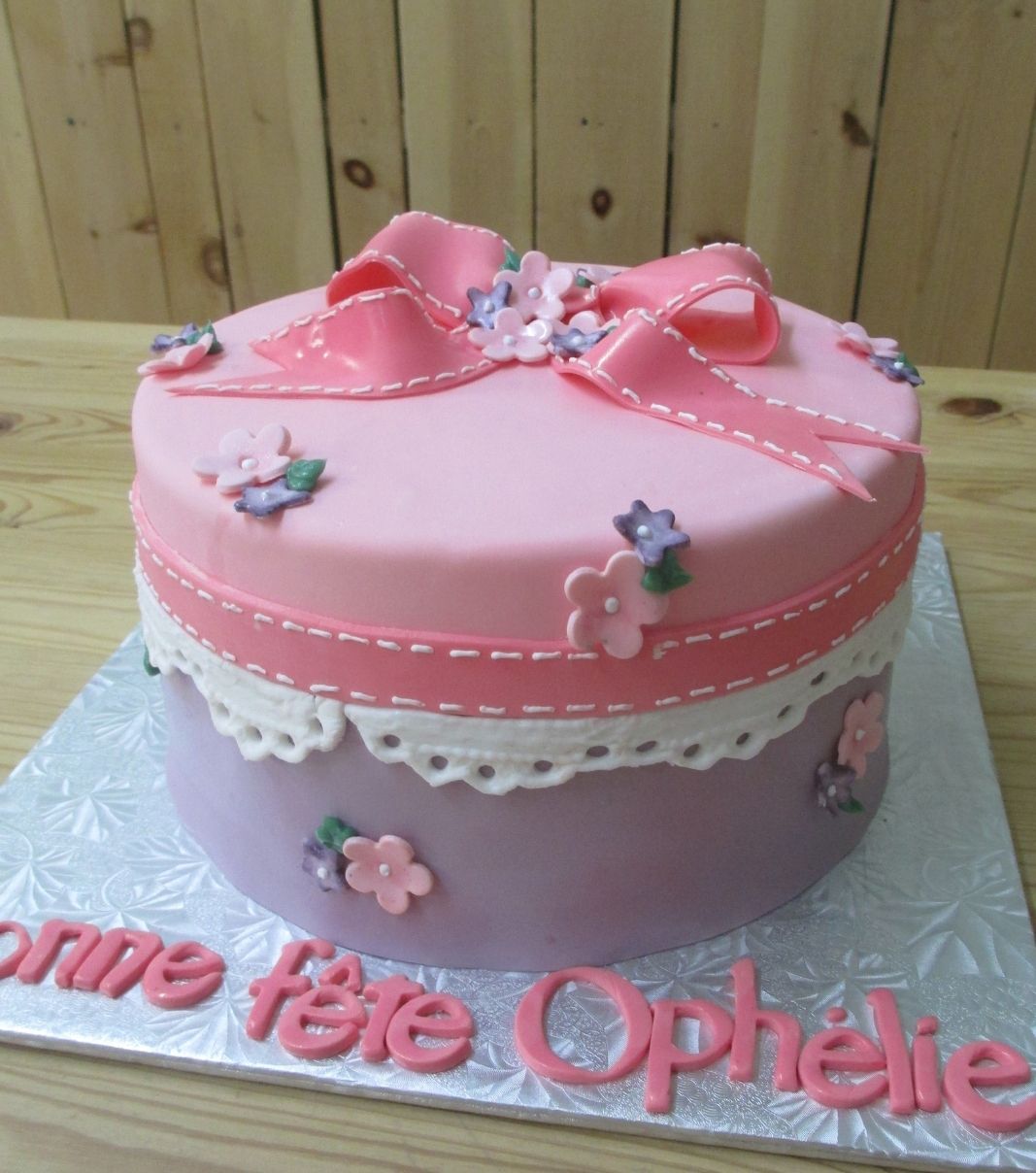 Gâteau d'anniversaire sur mesure d'Ophélie: gâteau avec une couverture en fondant à effet tissu mauve et rose et dentelle blanche décoré d'un ruban et de fleurs en fondant.