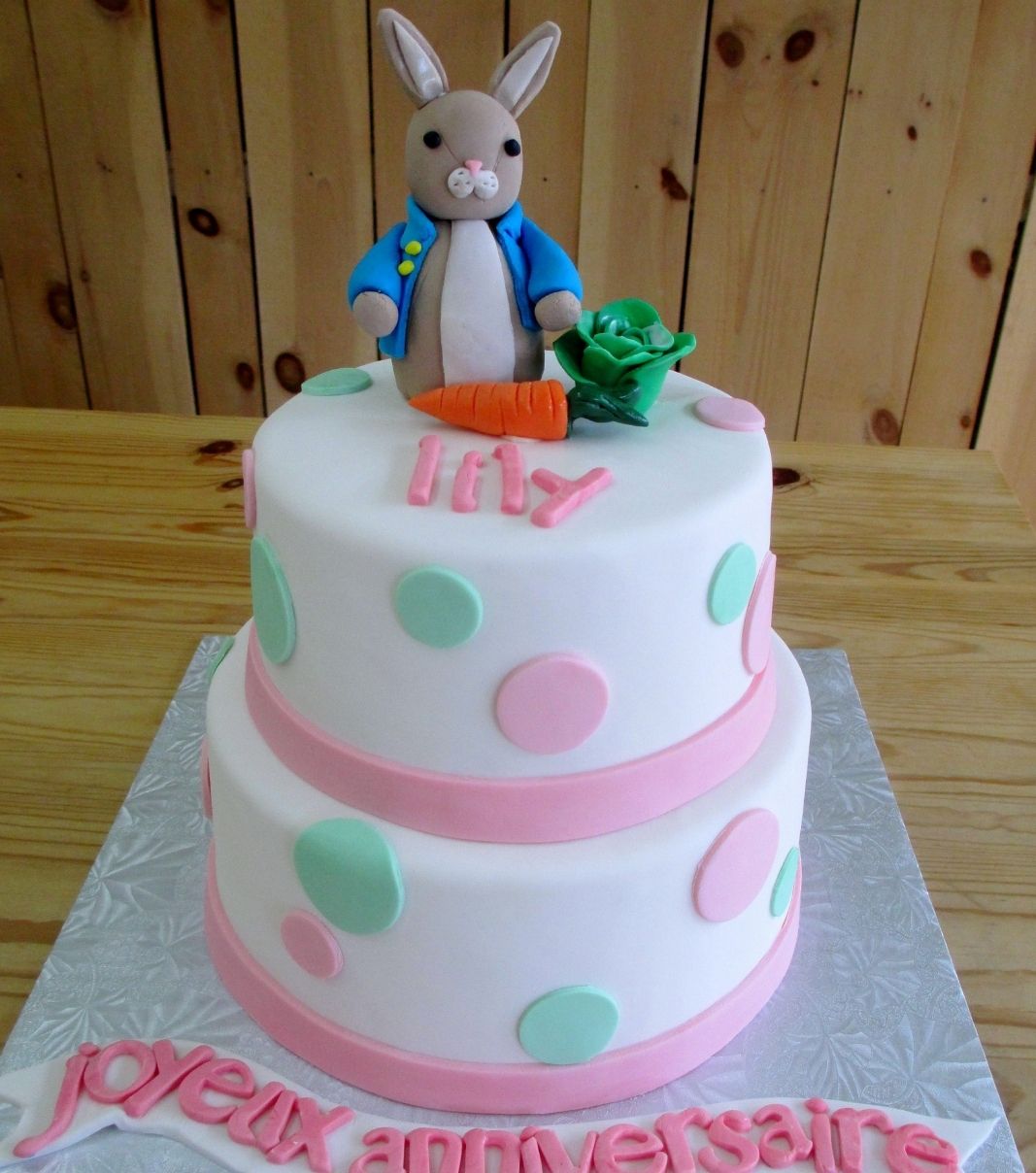 Gâteau d'anniversaire sur mesure de Lily: gâteau de 2 étages à thématique de Jeannot Lapin avec une couverture en fondant blanc et pastel et décoré d'un lapin et de légumes en fondant.