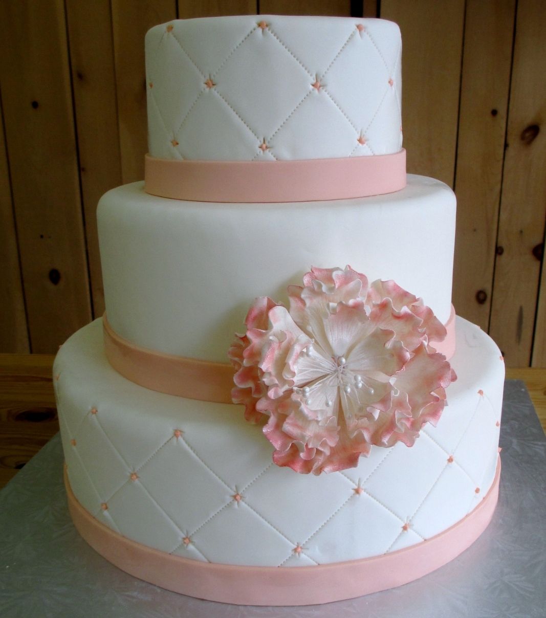 Gâteau de mariage: gâteau de 3 étages à thème blanc et pêche avec une couverture fondante à effet matelassé (quilting) et décoré d'une grosse fleur en fondant sucré avec une touche persillée de couleur rose pêche.