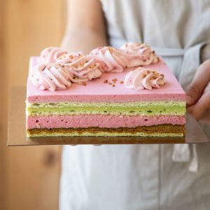 Photo du gâteau framboise pistache de Mlles Gâteaux : Mousse à la framboise sur croustillant à la pistache et un centre de crémeux à la pistache. Il est garni de chantilly à la framboise et de céréales croustillantes au chocolat blanc