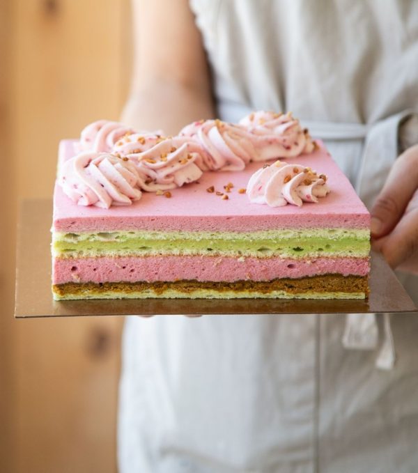 Photo du gâteau framboise pistache de Mlles Gâteaux : Mousse à la framboise sur croustillant à la pistache et un centre de crémeux à la pistache. Il est garni de chantilly à la framboise et de céréales croustillantes au chocolat blanc