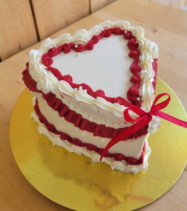 Gâteau en coeur style Lambeth. Saveur chocolat, vanille et cerise. Décor crème au beurre blanche et rouge avec boucle tissu rouge.
